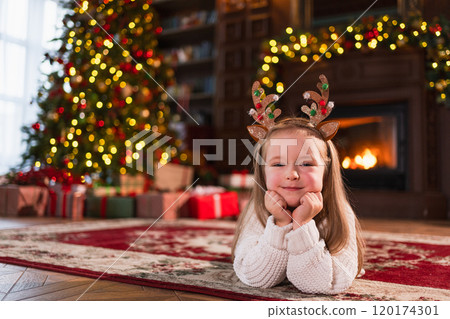 Merry Christmas. Little girl smiling near Christmas tree in classical dark interior. Young happy kid wearing deer horns in living room with fireplace Christmas tree gift boxes. Christmas eve at home 120174301