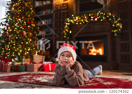 Merry Christmas. Little boy smiling near Christmas tree in classical dark interior. Young happy kid wearing Santa Hat in living room with fireplace Christmas tree gift boxes. Christmas eve at home 120174302