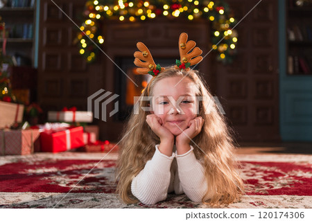 Merry Christmas. Little girl smiling near Christmas tree in classical dark interior. Young happy kid wearing deer horns in living room with fireplace Christmas tree gift boxes. Christmas eve at home 120174306