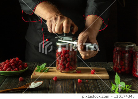 A cook rolls ripe raspberries in a jar with a hand seaming key. The concept of making canned sweet drink or jam on the kitchen table A cook rolls ripe raspberries in a jar with a hand seaming key. The concept of making canned sweet drink or jam on the kitchen table 120174343
