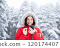 Beautiful woman in red hooded jacket, with phone in hands standing during day in winter snowy forest 120174407