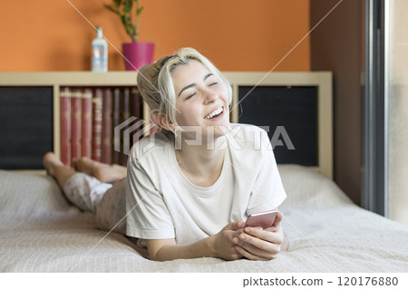 Laughing woman lying on bed, holding phone, indoor setting. Laughing woman lying on bed, holding phone, indoor setting. 120176880
