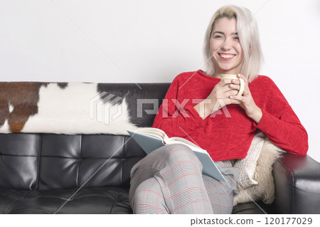 Smiling woman in red sweater with a mug and book on a couch. 120177029