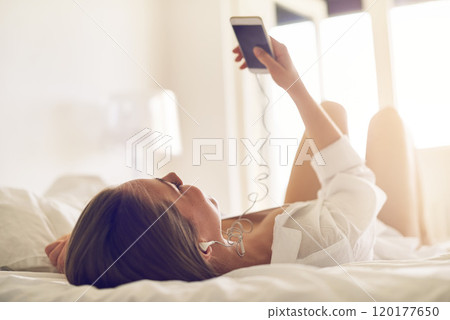 Beats to begin the day with. Shot of a young woman listening to music on her phone while relaxing on her bed. Beats to begin the day with. Shot of a young woman listening to music on her phone while relaxing on her bed. 120177650