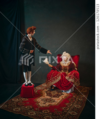 Young man, court servant in red wig standing next to elderly woman, medieval empress and singing to her. 120178513