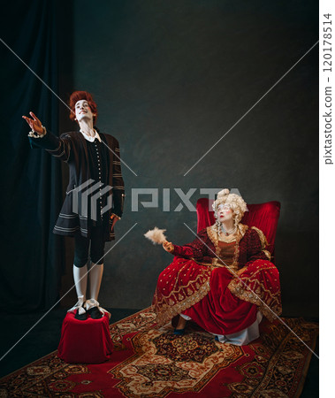 Young man, court servant in red wig standing next to elderly woman, medieval empress and singing to her. Young man, court servant in red wig standing next to elderly woman, medieval empress and singing to her. 120178514