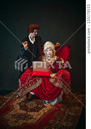 Impressed medieval elderly woman, royal empress sitting with laptop, young smiling man, court servant standing behind. Online shopping. 120178515