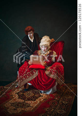 Impressed medieval elderly woman, royal empress looking on laptop with shocked face with young man, court servant standing behind 120178516