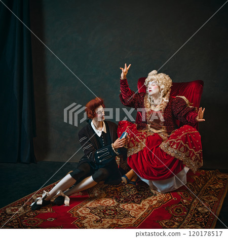 Elderly woman and young man against dark background. Medieval empress sitting in chair with young male servant holding her music player. 120178517