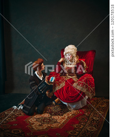 Humorous scene with elderly woman and young man. Medieval empress sitting in chair with young male servant offering her a music player. 120178518