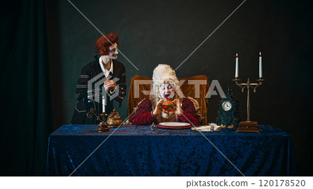 Young man, royal servant in wig, standing next to elderly woman, empress emotionally eating hamburger, against dark vintage background. Young man, royal servant in wig, standing next to elderly woman, empress emotionally eating hamburger, against dark vintage background. 120178520