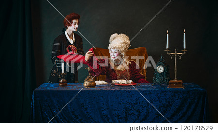 Loyal court favorite, young man presenting red luxury shoes to elderly woman. Royal empress cheerfully accepting gift 120178529