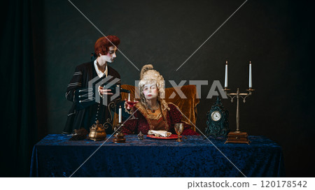 Young man, court servant pouring red wine to serious elderly woman, medieval empress with wig and weird makeup, sitting at table 120178542