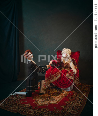 Empress from 18th century offering her hand to her favorite, who kneels in formal gesture of loyalty. Regal and historical charm 120178551