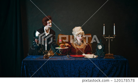 Young man, royal servant in wig, serving delicious hamburger to elderly woman, empress with serious face, set against dark vintage background. 120178578