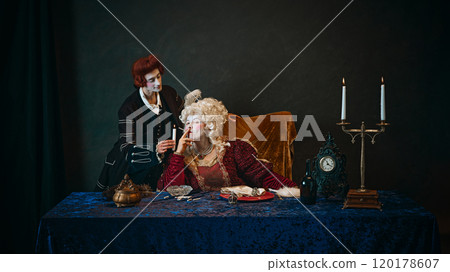 Royal empress, adorned in baroque gown, holding cigarette as her loyal favorite standing beside with candle. Royal empress, adorned in baroque gown, holding cigarette as her loyal favorite standing beside with candle. 120178607