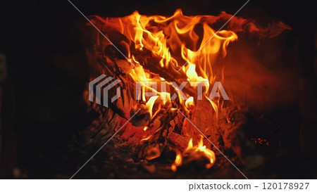 Cozy fireplace at night, burning wood logs glow warmly in shades of orange, red, and yellow, crackling and radiating heat 120178927