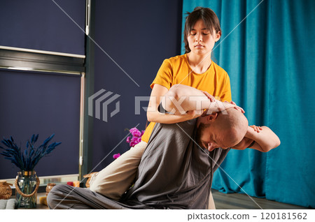 Masseuse making thai yoga massage. Woman therapist doing traditional massage treatment, stretching male patient's body. Bald client enjoying hand massage in calm atmosphere. 120181562