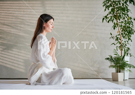 Beautiful woman in white dress sitting on mat in lotus position before Thai massage, practicing yoga, showing namaste sign. Beautiful woman in white dress sitting on mat in lotus position before Thai massage, practicing yoga, showing namaste sign. 120181565
