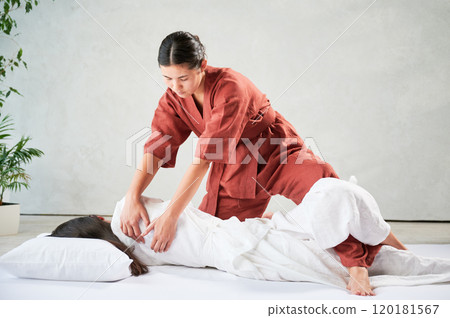Masseuse making thai yoga massage. Woman therapist doing traditional massage treatment, stretching female patient's body. Client lying on mat, enjoying hand massage in calm atmosphere. Masseuse making thai yoga massage. Woman therapist doing traditional massage treatment, stretching female patient's body. Client lying on mat, enjoying hand massage in calm atmosphere. 120181567