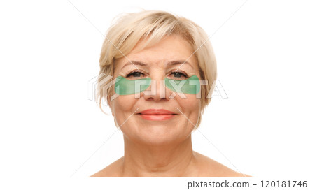 Close-up shot of senior blonde woman with green under-eye patches, looking directly at camera with a gentle smile against white studio background. 120181746