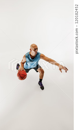 Male basketball player dressed in sports gear dribbles ball, focusing on his next move against grey studio background. Male basketball player dressed in sports gear dribbles ball, focusing on his next move against grey studio background. 120182452