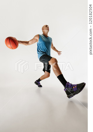 Low angle shot of male basketball player in athletic outfit dribbles, looking focused as he prepares to take his shot against grey studio background. 120182504