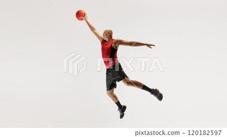 Young man in athletic clothing, wearing tank top and shorts, jumps with basketball toward hoop against grey studio background. 120182597