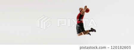 Male athlete dressed in tank top and athletic shorts leaps high, aiming to dunk basketball against grey studio background. 120182603