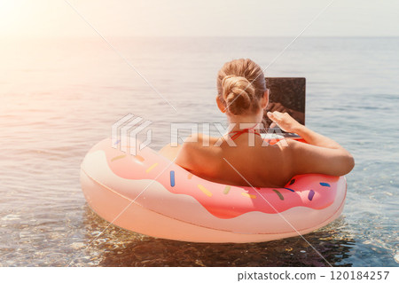 Woman Laptop Beach - A woman sits on a donut floatie in the ocean, working on her laptop. The sun shines brightly in the background. 120184257