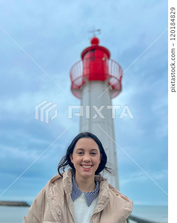 Woman standing on the wooden pier and looking at the red lighthouse. Red lighthouse with wooden pier with blue sky with rainy clouds background in the Normandia, France. 120184829