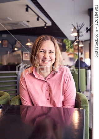 Smiling woman in pink shirt relaxing at modern cafe with cozy interior and green Smiling woman in pink shirt relaxing at modern cafe with cozy interior and green 120184903