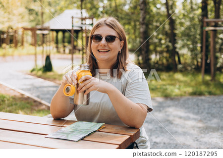 Happy 30s Woman in active trekking clothes having a halt after hiking. Hiker drinking water from water bottle or hot drink from yellow thermos. Woman planning new travel with paper map 120187295