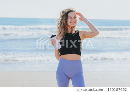 Fitness, sport and healthy active lifestyle. Happy slim young athlete lady in sportswear holds bottle of water, rests after workout on sea beach. Break, hydration, wellness, healthcare, aqua balance 120187662