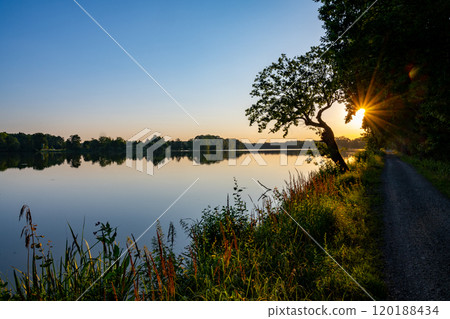 The tranquil Trebonsko Pond System in South Bohemia glows as the sun sets, reflecting golden light on calm waters flanked by lush greenery and a peaceful pathway. 120188434