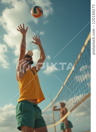 A man in a yellow tank top and green shorts is jumping to set a volleyball against a bright blue sky with fluffy clouds. The scene captures the energy and excitement of beach volleyball, showcasing 120189275