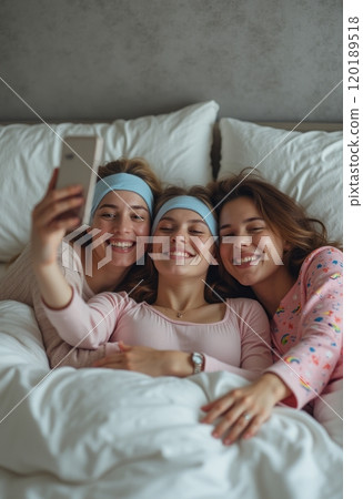 Three young women are lying together in a cozy bed, smiling and taking a selfie with a smartphone. They are wearing matching headbands and comfortable pajamas, creating a warm and joyful atmosphere of 120189518
