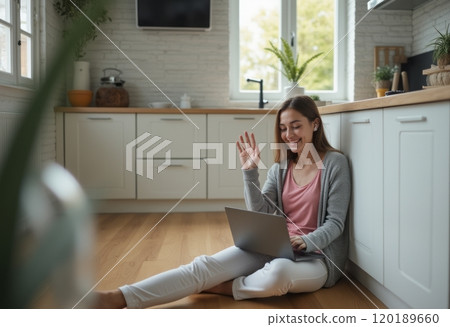 A young woman is sitting on the kitchen floor, smiling and waving at her laptop while engaging in a video call. The bright and airy kitchen features natural light streaming through the windows 120189660