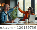 A man and a woman are joyfully giving each other a high five in a modern office setting, showcasing teamwork and enthusiasm. The woman, wearing an orange blazer, is smiling broadly, while the man, in 120189736