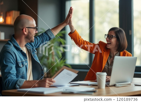 A man and a woman are joyfully giving each other a high five in a modern office setting, showcasing teamwork and enthusiasm. The woman, wearing an orange blazer, is smiling broadly, while the man, in 120189736