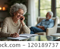 An elderly woman with curly gray hair is smiling while talking on the phone, sitting at a table with a notebook in front of her. In the background, an older man is seated comfortably, creating a warm 120189737
