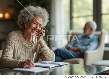 An elderly woman with curly gray hair is smiling while talking on the phone, sitting at a table with a notebook in front of her. In the background, an older man is seated comfortably, creating a warm 120189737