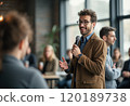 A confident man with curly hair and glasses is speaking into a microphone while gesturing expressively in a modern indoor setting. The audience, partially visible, appears engaged and attentive 120189738