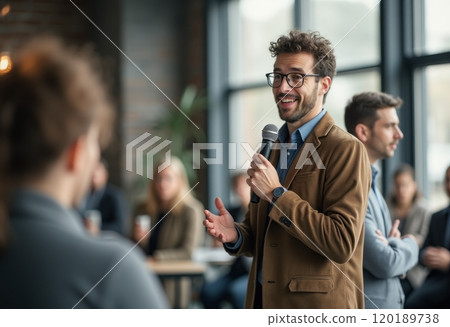 A confident man with curly hair and glasses is speaking into a microphone while gesturing expressively in a modern indoor setting. The audience, partially visible, appears engaged and attentive 120189738