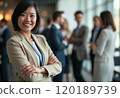 A confident businesswoman stands with her arms crossed, smiling warmly at the camera while a group of professionals engages in conversation in the background. The image captures a dynamic business 120189739