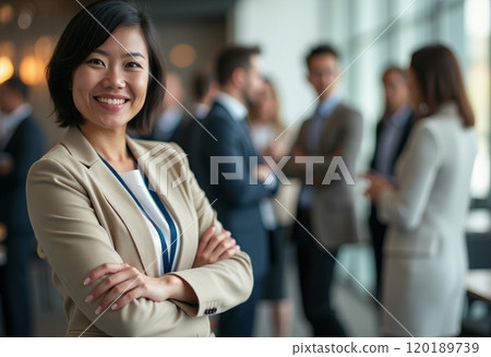 A confident businesswoman stands with her arms crossed, smiling warmly at the camera while a group of professionals engages in conversation in the background. The image captures a dynamic business 120189739