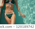 A woman in a green bikini is lounging by the edge of a sparkling pool, with her hand gently touching the water. The image captures a serene summer vibe, highlighting the beauty of relaxation and the 120189742