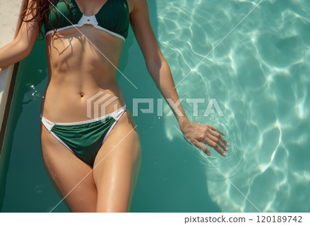 A woman in a green bikini is lounging by the edge of a sparkling pool, with her hand gently touching the water. The image captures a serene summer vibe, highlighting the beauty of relaxation and the 120189742