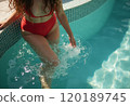 A woman in a vibrant red bikini is playfully splashing water while sitting on the edge of a pool. The scene captures a moment of relaxation and joy, highlighting the shimmering blue water and the 120189745