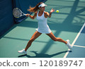 A focused female tennis player is captured mid-swing on a bright court, showcasing her athleticism and skill. She wears a white outfit and a visor, demonstrating a dynamic pose as she prepares to hit 120189747